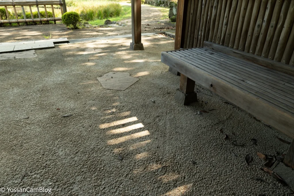 木陰のベンチと玉砂利の地面。16mm・1/13秒の手持ちで、砂利の粒まで止まっている